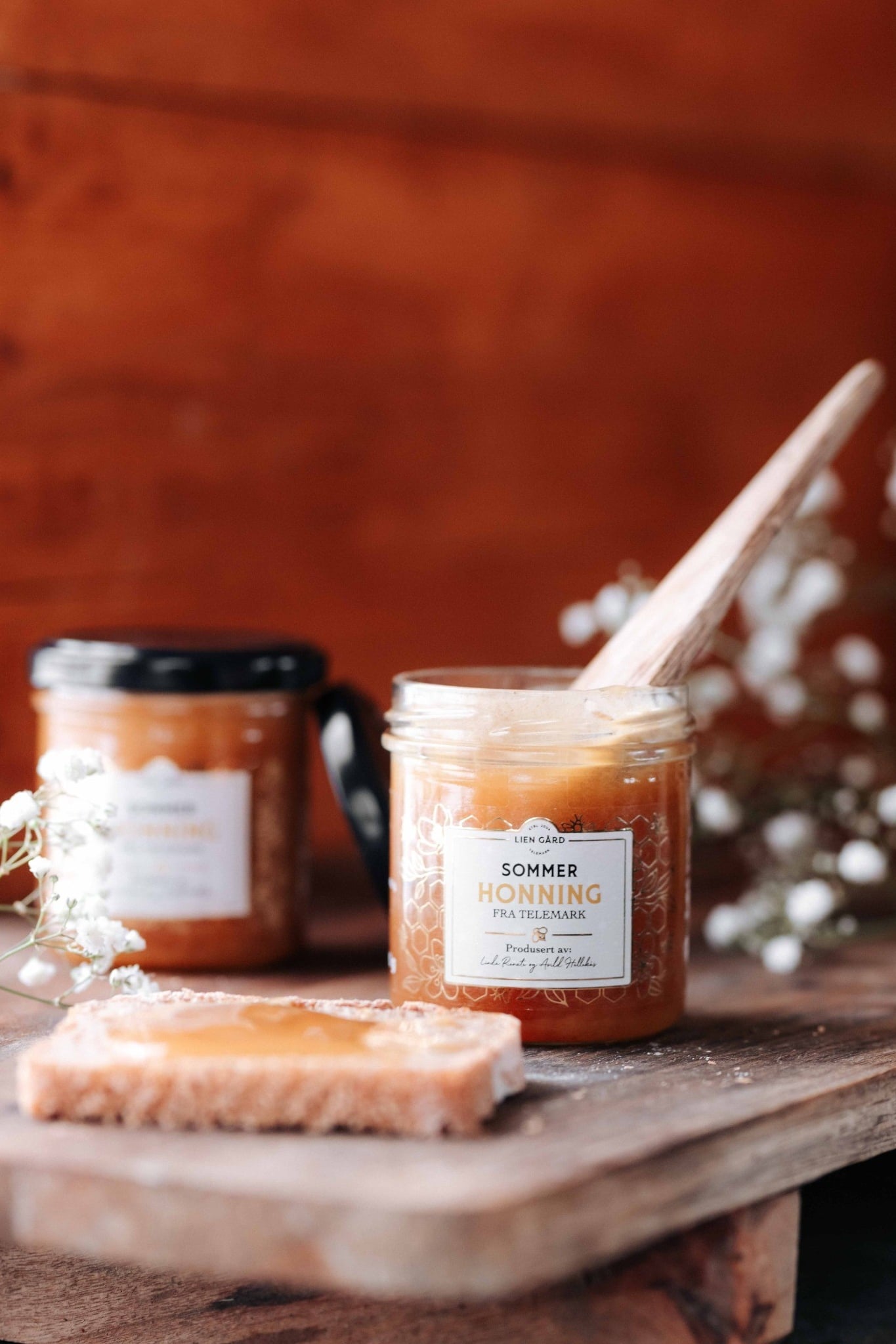Glas med sommerhonning fra Telemark, plassert på bord med brødskive og dekorative blomster i bakgrunnen.